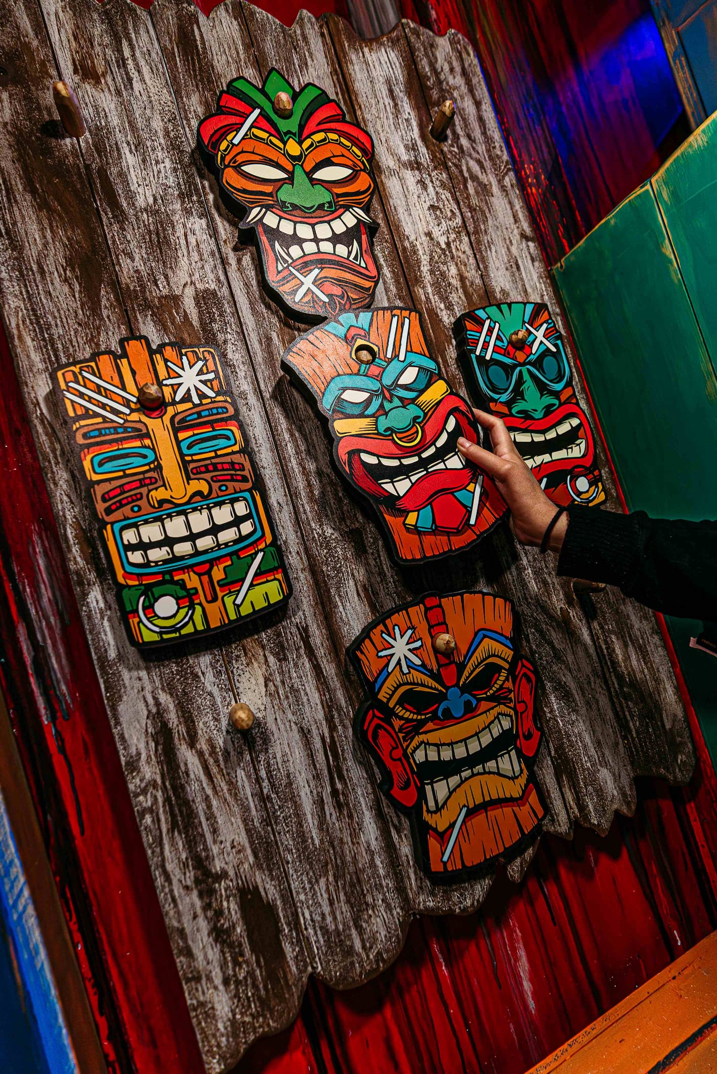 A hand reaches towards five colorful tiki masks mounted on a wooden wall.